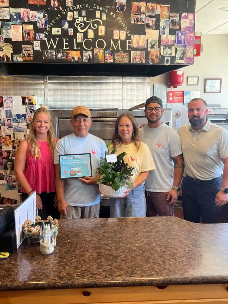 Winn and Sara behind their counter accepting an Iowa State Fair finalist certificate from fair presenters, under the restaurant's welcome sign